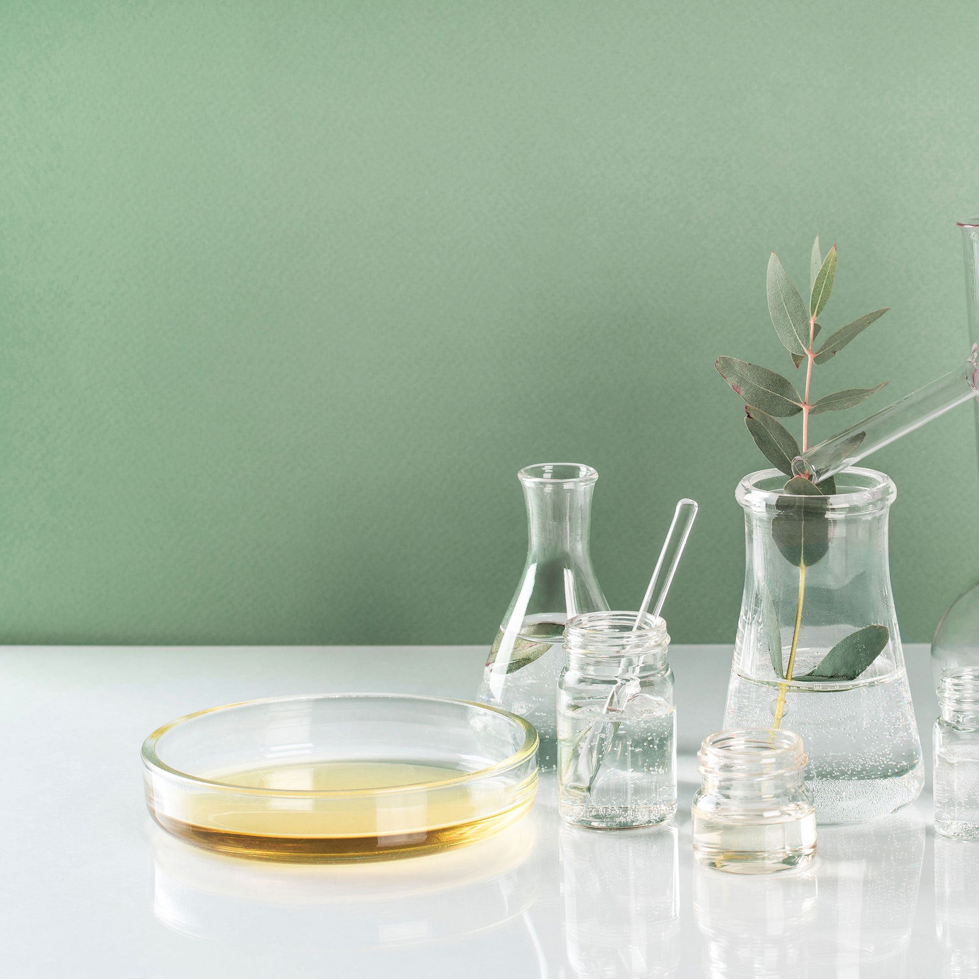 Bol en verre d’huile dorée et flacons avec feuilles et solution claire sur fond vert.