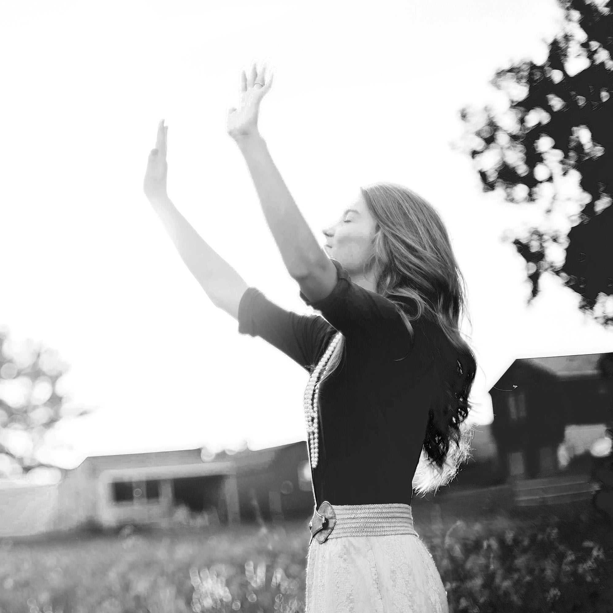 Femme debout en extérieur, bras levés vers le ciel, visage tourné vers la lumière.