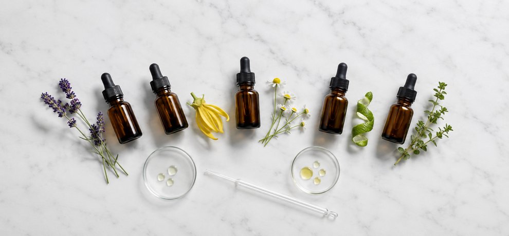 Cinq flacons ambrés d'Huiles Essentielles avec lavande, ylang ylang, camomille, bergamote et marjolaine sur surface blanche