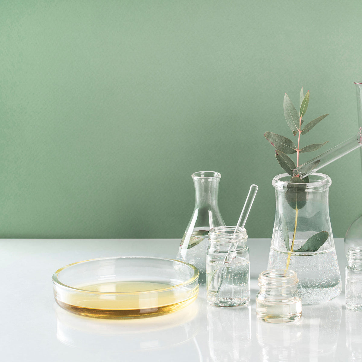 Bol en verre d’huile dorée et flacons avec feuilles et solution claire sur fond vert.